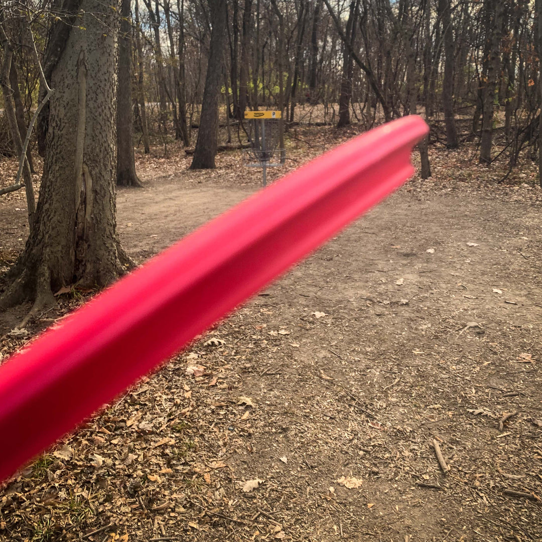 A red Prophecy - SureGrip overstable mid-range disc soars toward a disc golf basket through leaf-strewn, bare trees, highlighting its consistent flight in the woods.