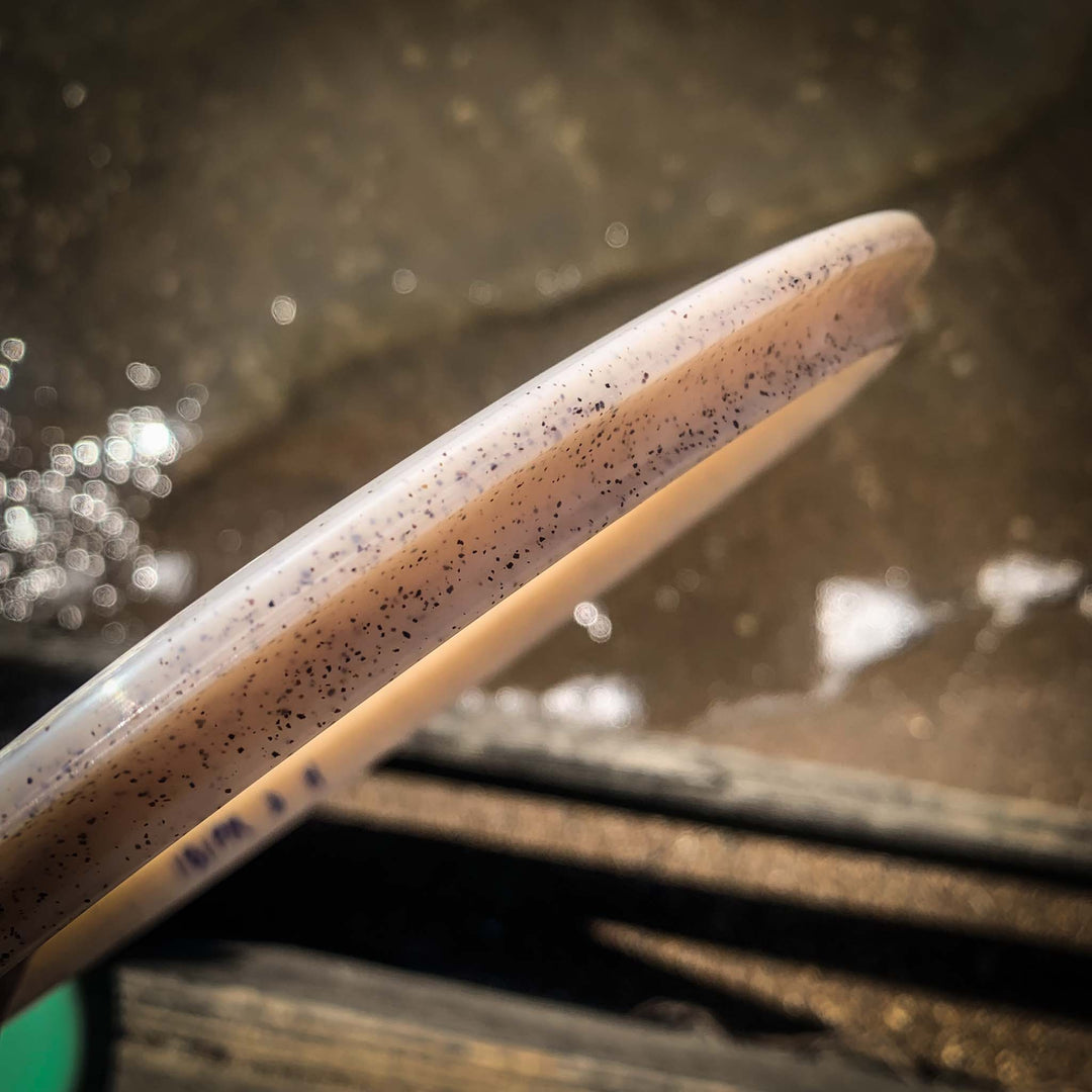 A close-up of the Prophecy - Diamond Hemp flying disc's rim, featuring small black speckles, held above a blurred water and dock background—ideal for consistent flight.