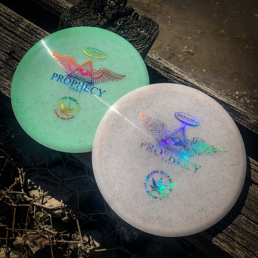 Two premium Prophecy - Diamond Hemp discs with holographic triangle and wing designs rest on the ground by wood and water, ready to deliver consistent flight with every throw.