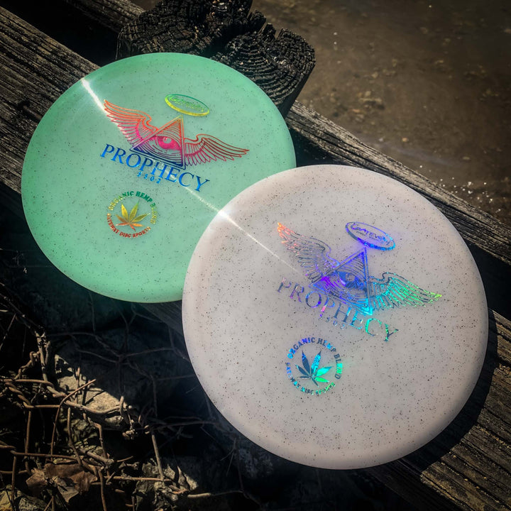 Two premium Prophecy - Diamond Hemp discs with holographic triangle and wing designs rest on the ground by wood and water, ready to deliver consistent flight with every throw.