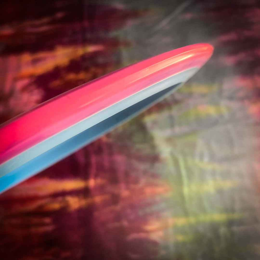 A close-up of the rim of the Prototype 93 disc golf disc in pink, white, and blue showcases its dual-material technology, with a blurred multicolored background highlighting its precision and innovation.