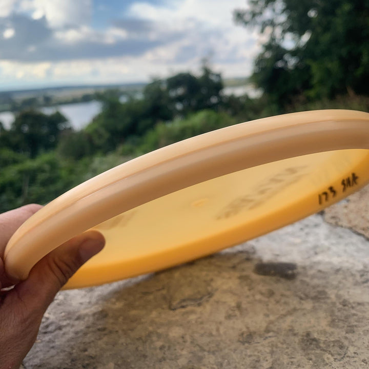 A hand holds a yellow Shaman - Diamond disc golf disc outdoors near a stone ledge, surrounded by trees, water, and a cloudy sky—ready to test its flight path in nature.