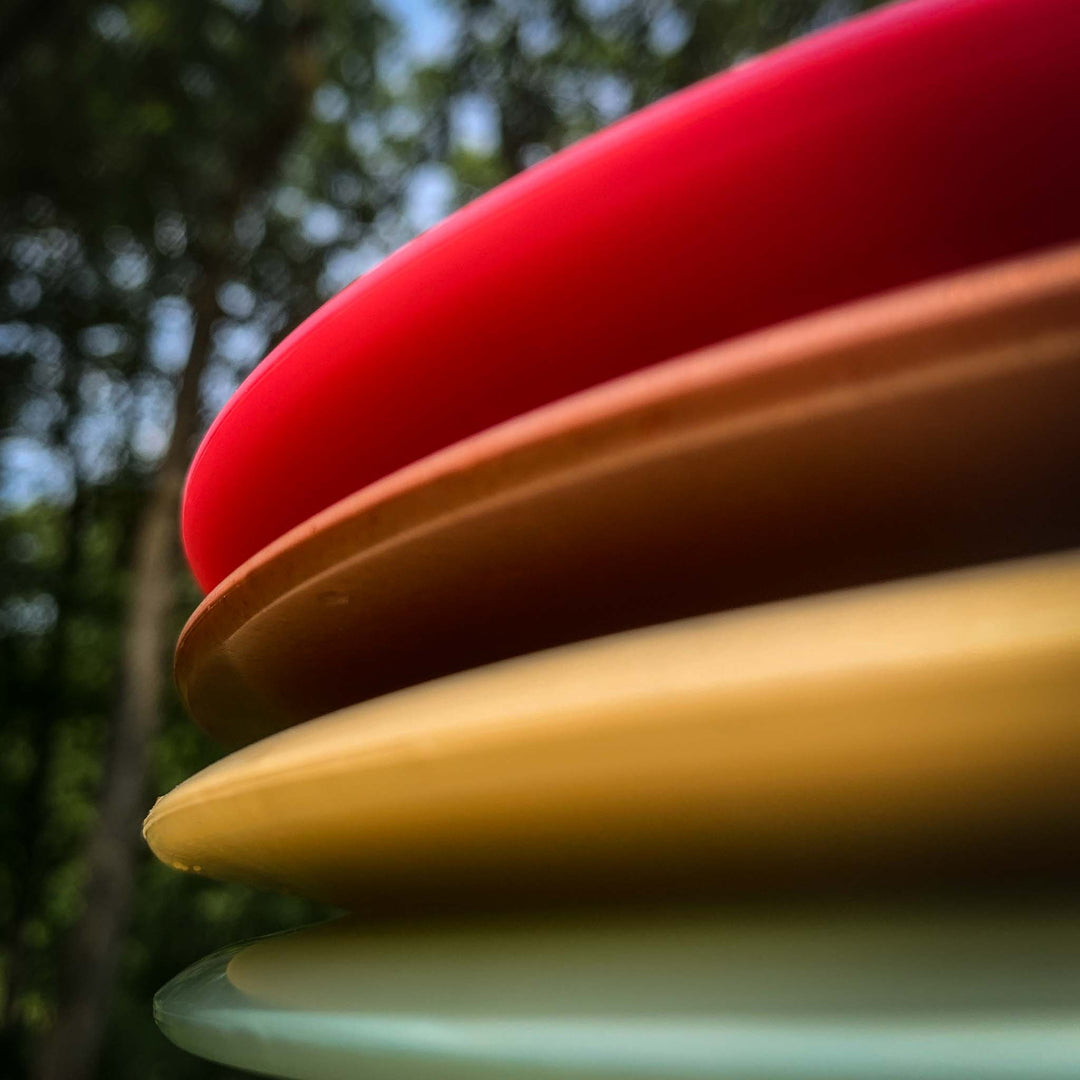 Close-up of the edges of four stacked Spear - Diamond plastic discs in red, orange, yellow, and white—highlighting a straight control driver—set against a blurred outdoor background.