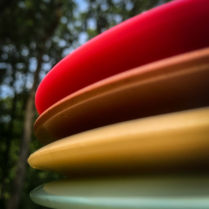 Close-up of the edges of four stacked Spear - Diamond plastic discs in red, orange, yellow, and white—highlighting a straight control driver—set against a blurred outdoor background.