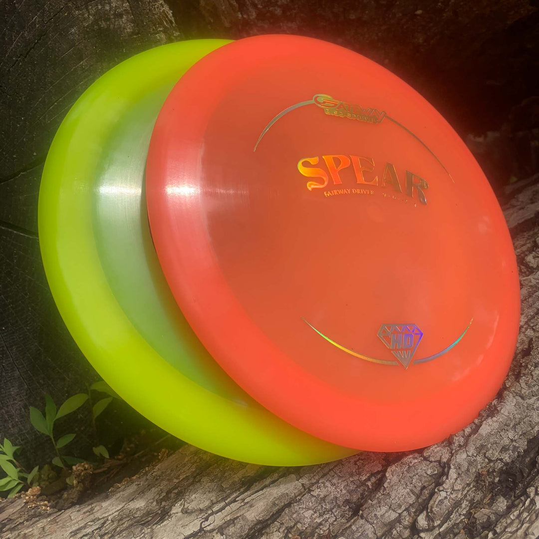 Two disc golf discs, one bright yellow and one bright orange labeled "Spear - Hyper Diamond (HD)," are stacked against a tree—perfect control drivers with a comfortable grip for smooth, accurate throws.