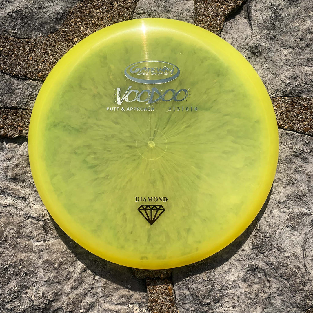 A Voodoo - Diamond disc golf putter in yellow, prized for its comfortable grip and straight-line flight, sits on a stone surface in sunlight.