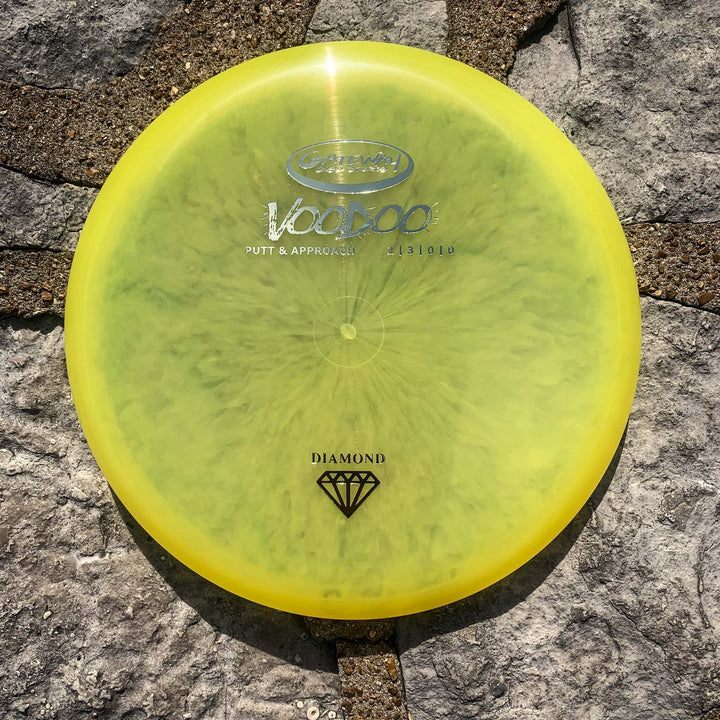 A Voodoo - Diamond disc golf putter in yellow, prized for its comfortable grip and straight-line flight, sits on a stone surface in sunlight.