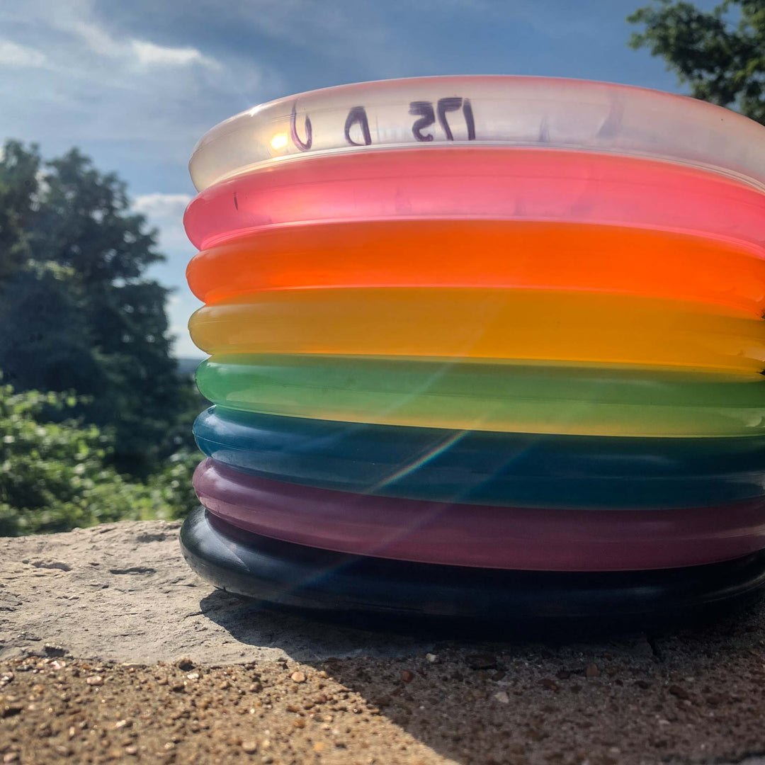 The Voodoo - Diamond, a vibrant stack of translucent discs with a comfortable lay-up disc on top, sits on a stone surface outdoors, surrounded by greenery and blue sky.