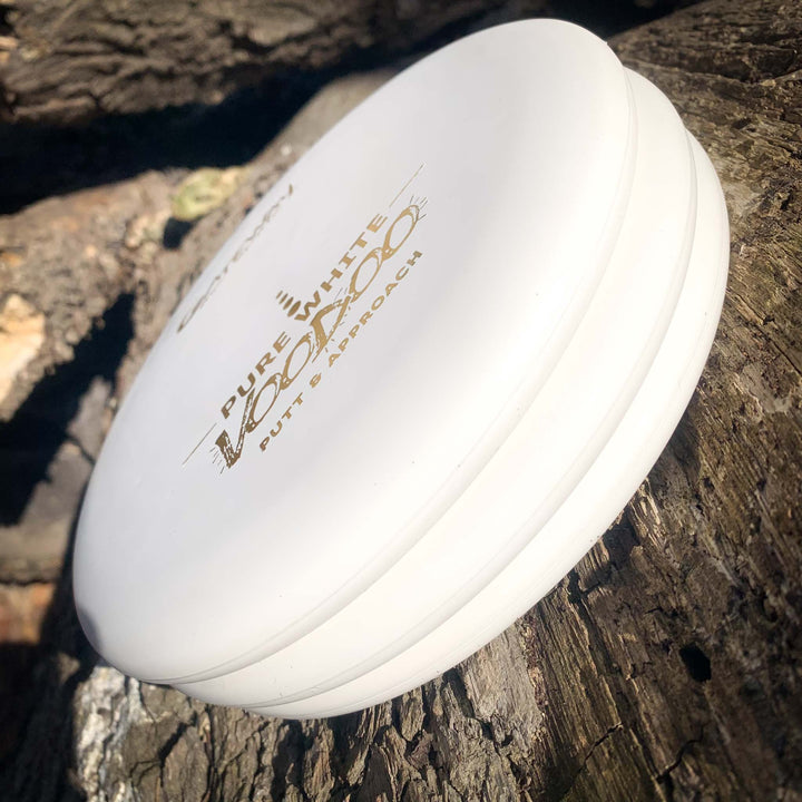 A white Voodoo - Pure White putter rests on a rough tree trunk in sunlight.