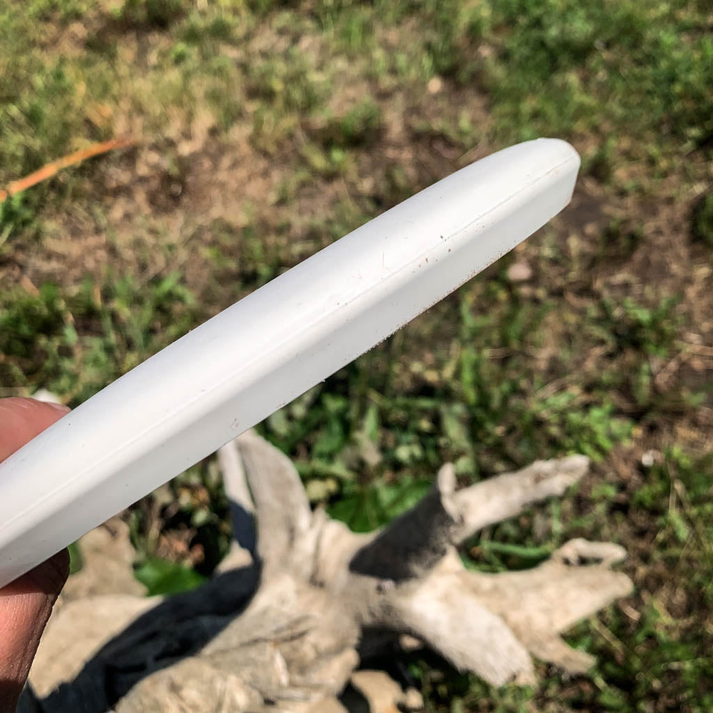 Someone holds a white Warlock - Suregrip - SSSS disc-shaped putter outdoors, with grass and a piece of driftwood visible in the background.
