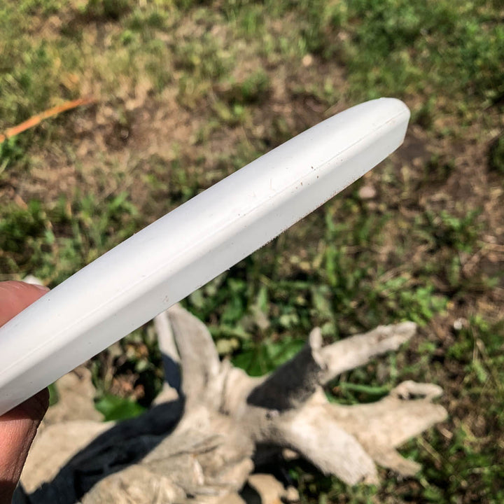 Someone holds a white Warlock - Suregrip - SSSS disc-shaped putter outdoors, with grass and a piece of driftwood visible in the background.