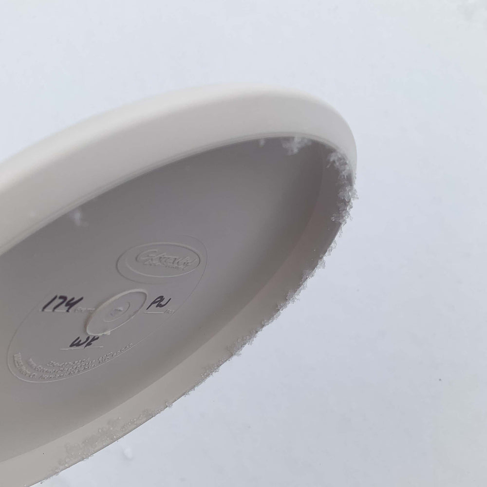 A close-up of the underside of a Warlock - Pure White disc with handwritten markings and a thin layer of frost along the edge, set against a snowy background.