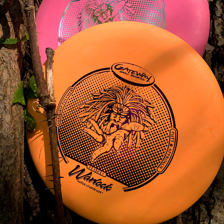 An orange Warlock - Suregrip - SSS disc golf putter featuring a person with dreadlocks leans against a tree, with a pink disc partly visible behind it.