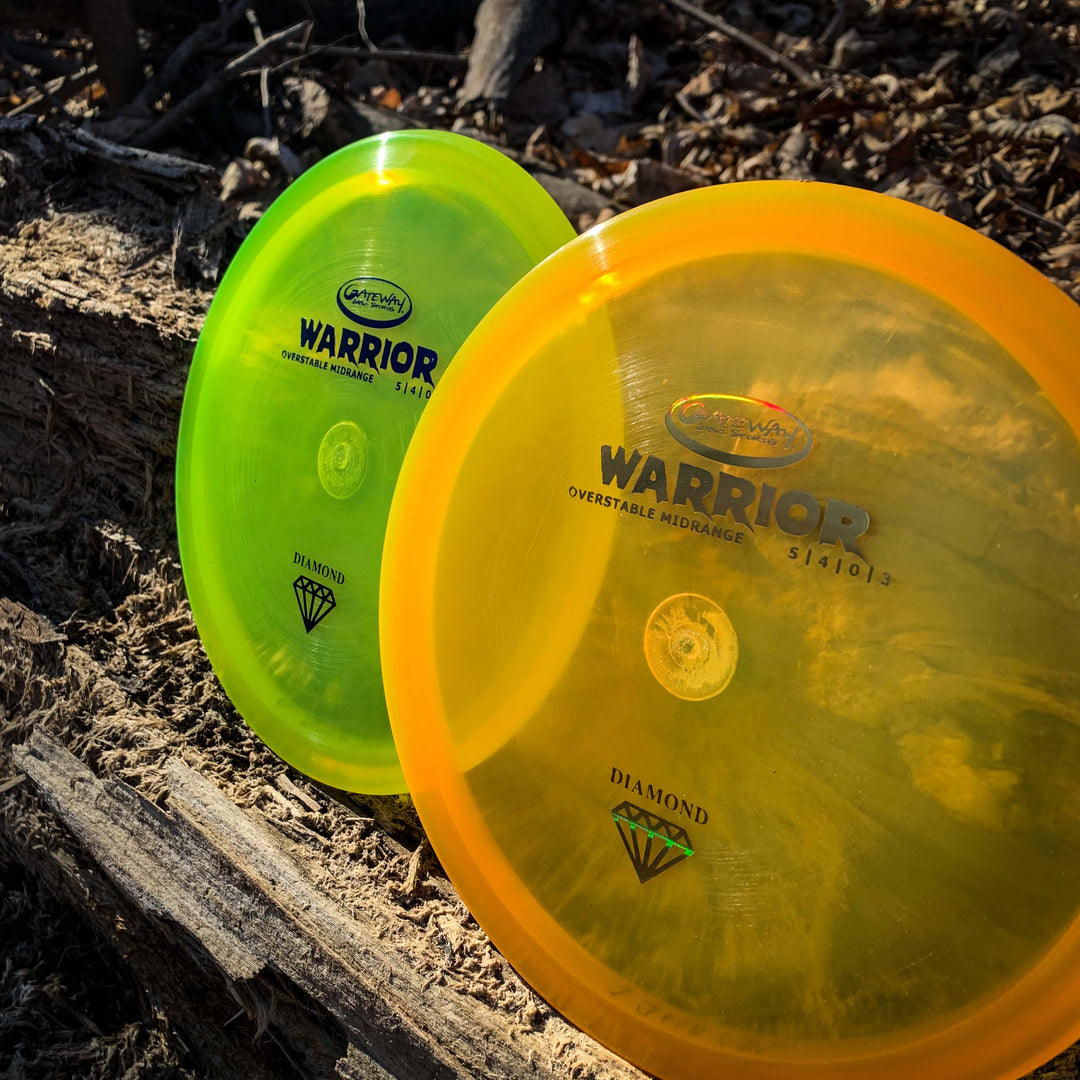 Two overstable Warrior - Diamond mid-range disc golf discs, one yellow-green and one orange, stand upright against logs and twigs on a forest floor.