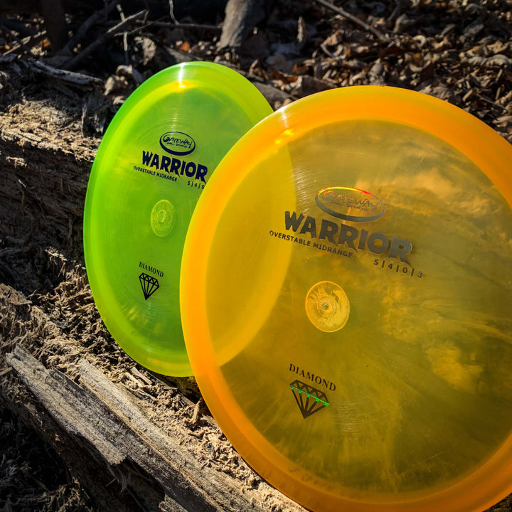 Two overstable Warrior - Diamond mid-range disc golf discs, one yellow-green and one orange, stand upright against logs and twigs on a forest floor.
