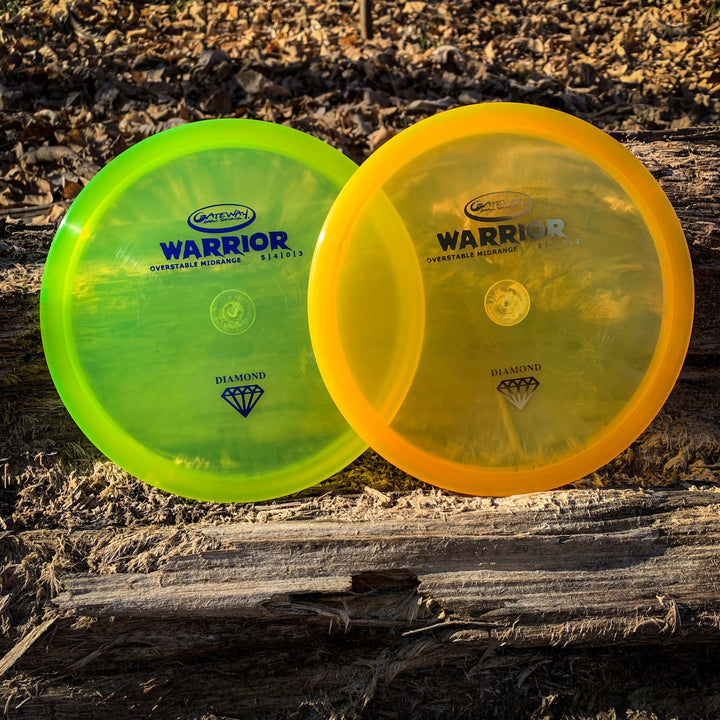 Two overstable "Warrior - Diamond" mid-range discs by Gateway, one yellow-green and one orange, rest on a log surrounded by wood chips and debris.