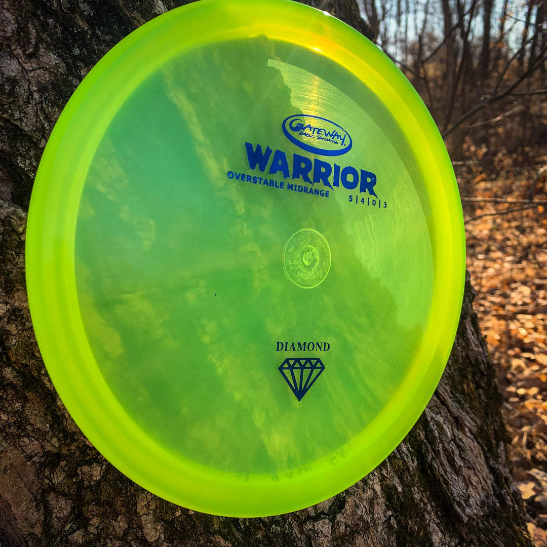 A yellow-green Warrior - Diamond overstable mid-range disc golf disc leans against a tree trunk in a wooded area with scattered fallen leaves.