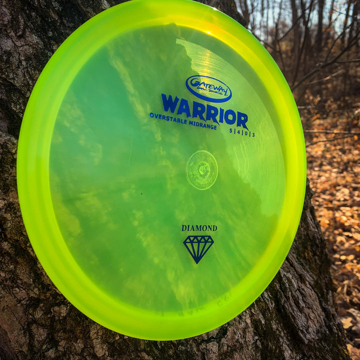 A yellow-green Warrior - Diamond overstable mid-range disc golf disc leans against a tree trunk in a wooded area with scattered fallen leaves.