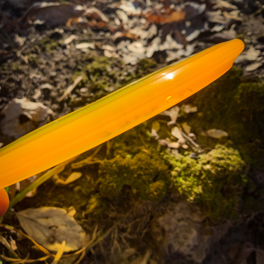 A close-up side view of the Warrior - Diamond mid-range disc in yellow-orange, held outdoors above mossy ground and dry leaves in sunlight.