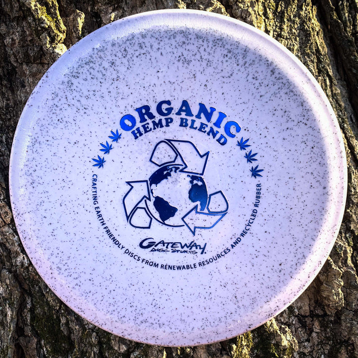 A Warrior - Organic Hemp mid-range disc golf disc, white with blue "Organic Hemp Blend" text and recycling symbols, leans against a tree trunk. This overstable disc features eco-friendly materials for sustainable play.