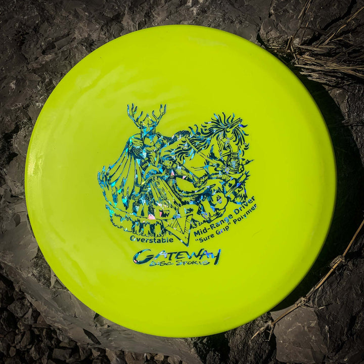 A yellow Gateway Warrior - SureGrip overstable mid-range disc with blue artwork, labeled "Cverstable Mid-Range Driver Sure Grip Polymer," ideal for long upshots, rests on rocky ground.