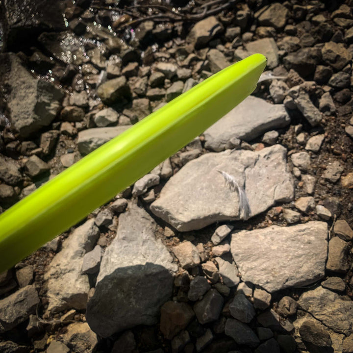 A Warrior - SureGrip disc in bright yellow-green is held above rocky ground, sunlight casting shadows—perfect for long upshots or when an overstable mid-range throw is needed.