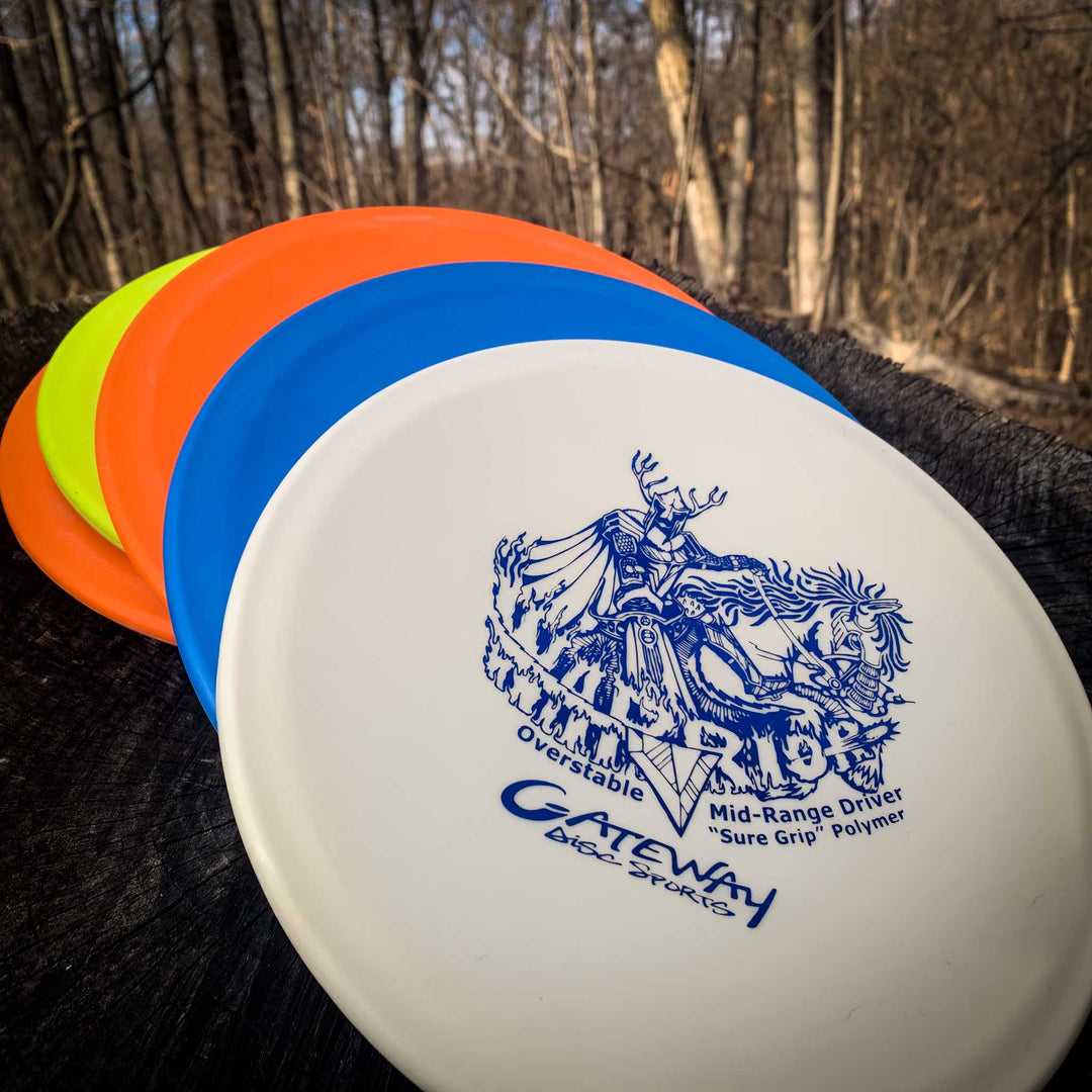 Four colorful disc golf discs, including a white Warrior - SureGrip overstable mid-range disc, are displayed in a fan shape on a tree stump in a wooded outdoor setting.