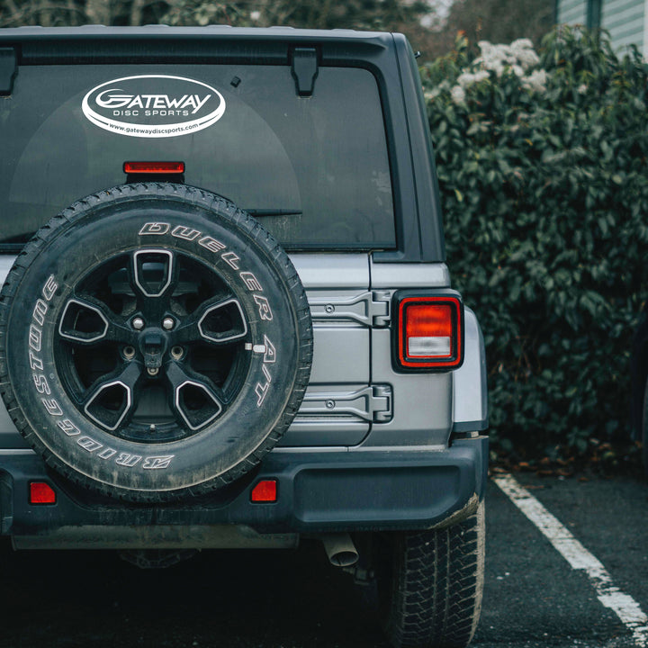 Rear view of a gray SUV with a white Gateway Disc Sports Team Window Decal on the back window and a rear-mounted spare tire, parked by green bushes. Great for adding style to your car windows.