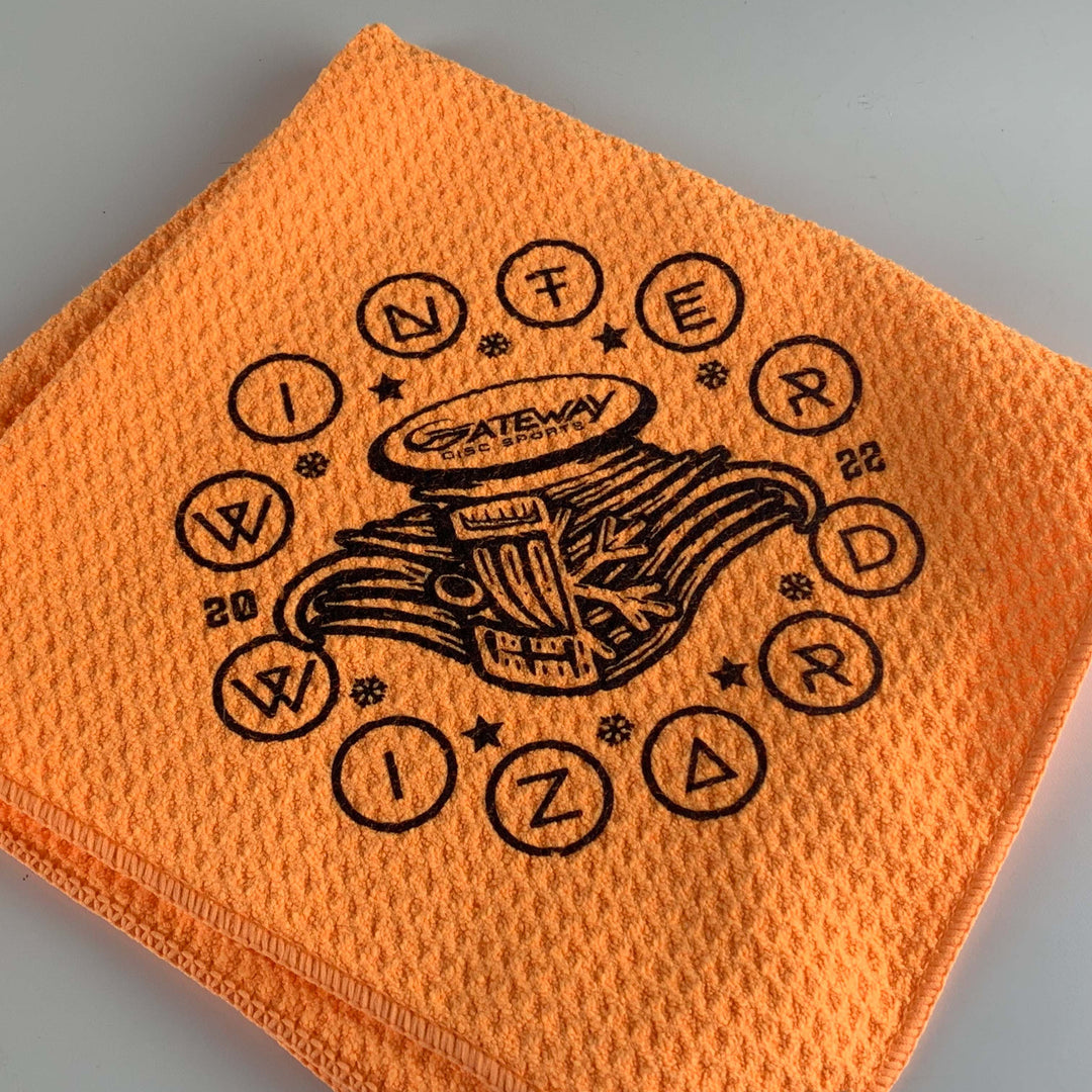 The Gateway Logoed Winter Wizards Towel is an orange cotton waffle towel with a black "Winter Wizard 2022" design and book stack, labeled "Gateway Disc Sports." Hemmed edges provide added durability and style.