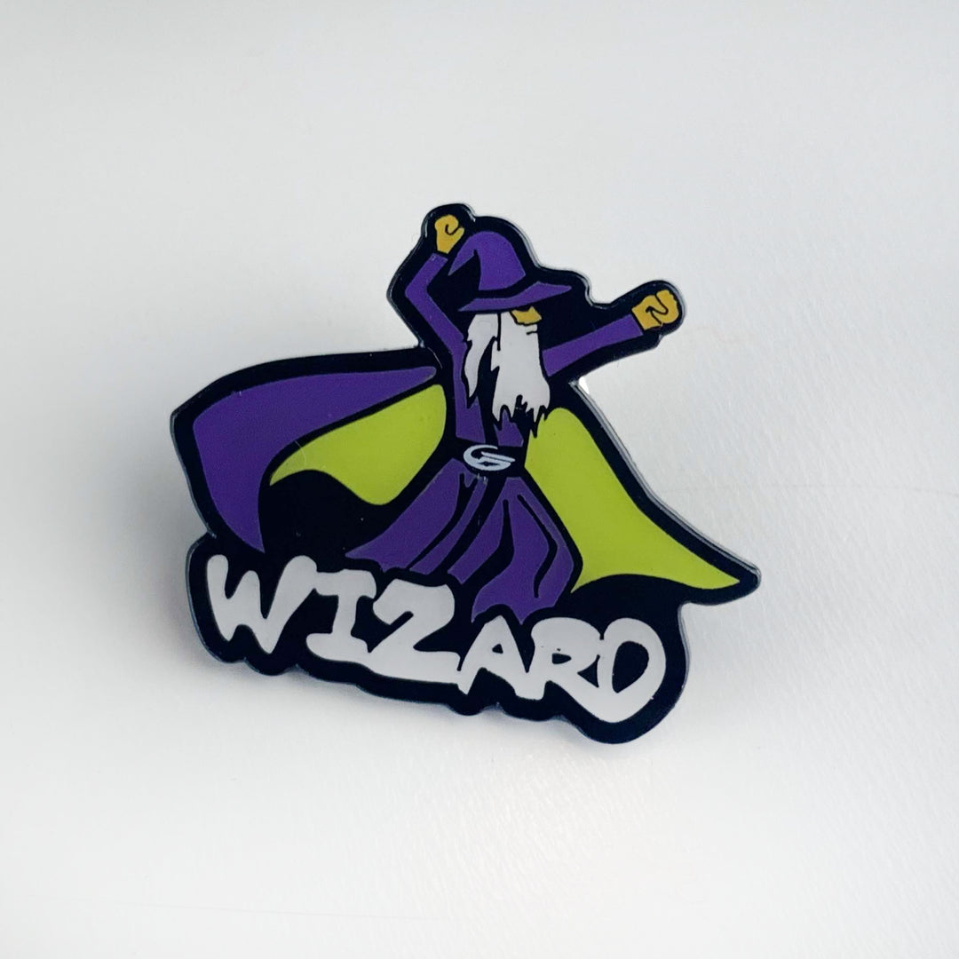 The Wizard Hat Pin is a Black Nichol pin featuring a cartoon wizard in a purple robe and hat with a yellow cape, arms raised above bold "WIZARD" text. It comes with a secure butterfly clutch for easy attachment.