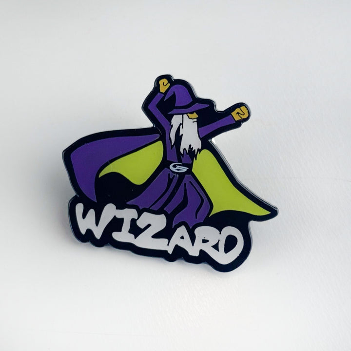 The Wizard Hat Pin is a Black Nichol pin featuring a cartoon wizard in a purple robe and hat with a yellow cape, arms raised above bold "WIZARD" text. It comes with a secure butterfly clutch for easy attachment.