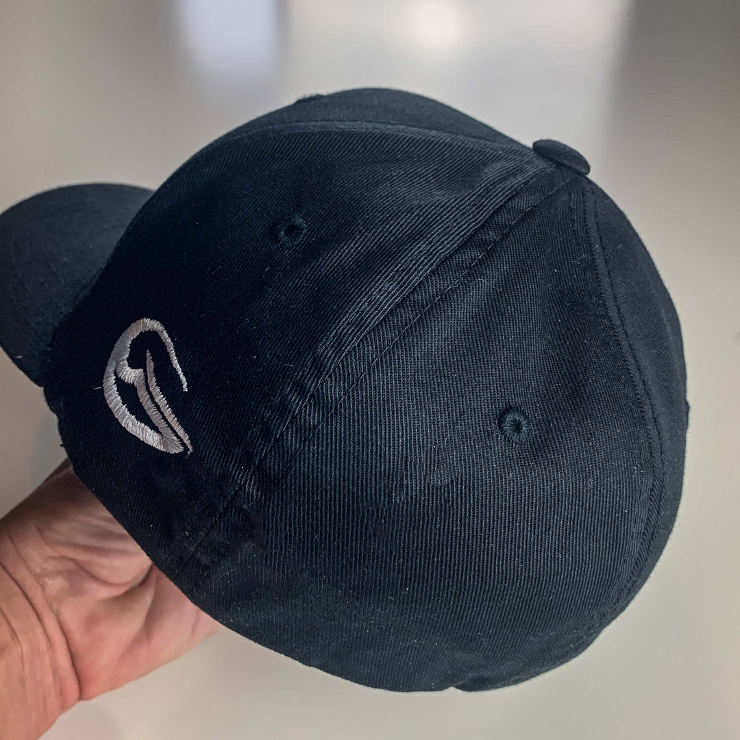 A hand holds a Wizard Flexfit Fitted Hat in black, featuring a silver embroidered Mini-Wizard logo on the side, set against a softly blurred background.