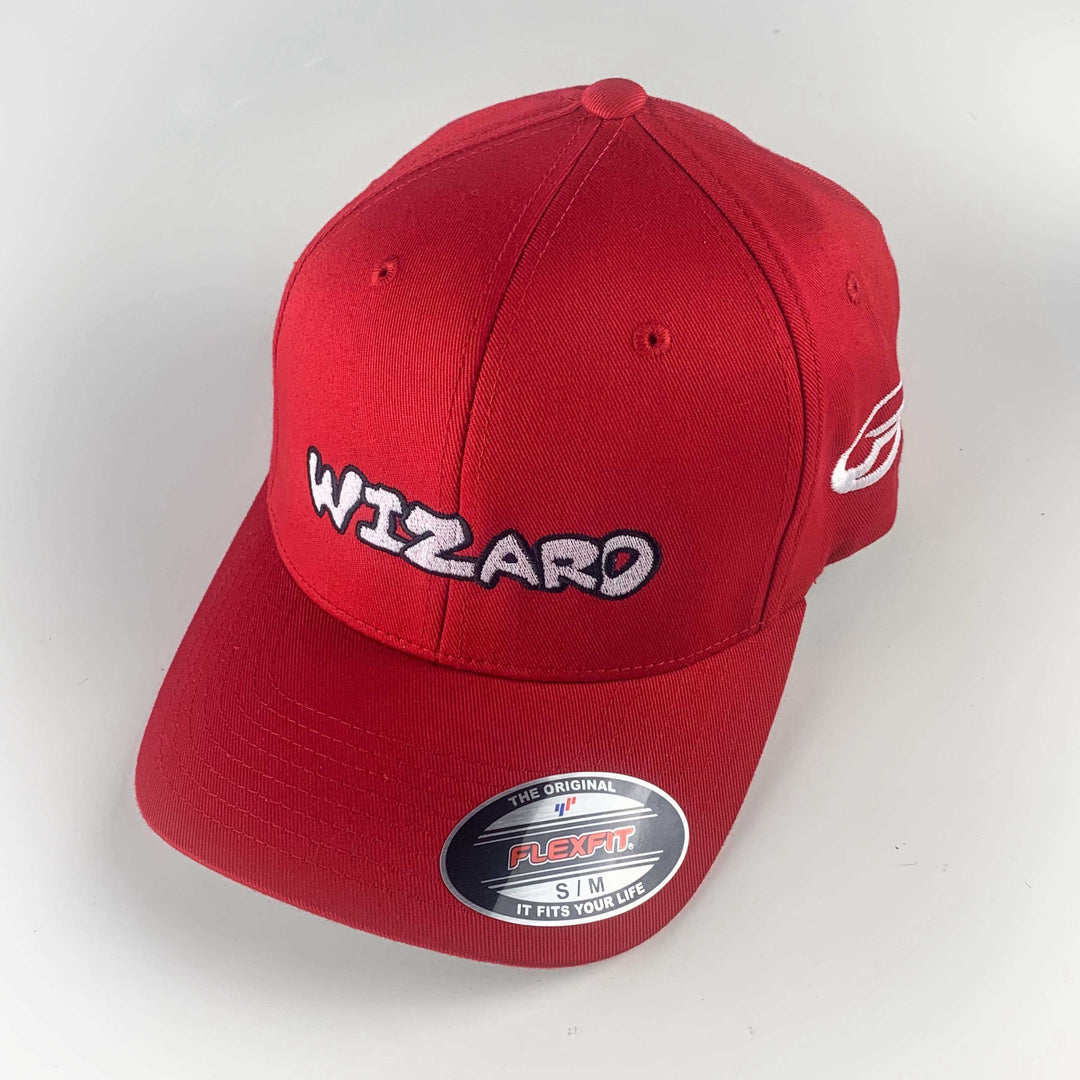 The Wizard Flexfit Fitted Hat features a red design, "WIZARD" embroidered in white and black on the front, a silver FLEXFIT sticker on the brim, and a subtle Mini-Wizard bar stamp detail.