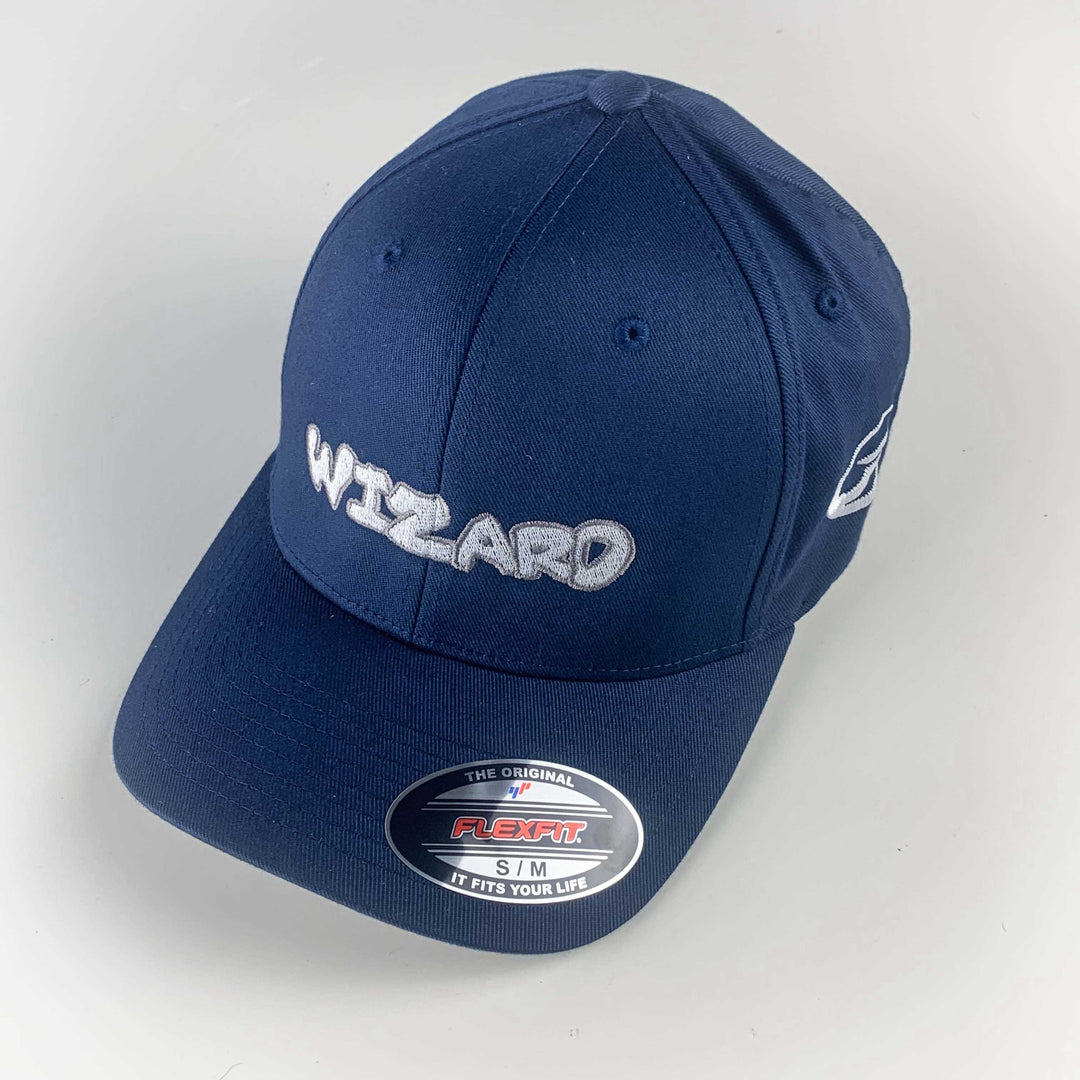 The Wizard Flexfit Fitted Hat features "WIZARD" embroidered in white on navy blue, a Flexfit sticker on the brim, and is inspired by the iconic Wizard bar stamp style.