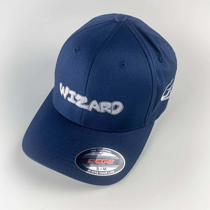 The Wizard Flexfit Fitted Hat features "WIZARD" embroidered in white on navy blue, a Flexfit sticker on the brim, and is inspired by the iconic Wizard bar stamp style.