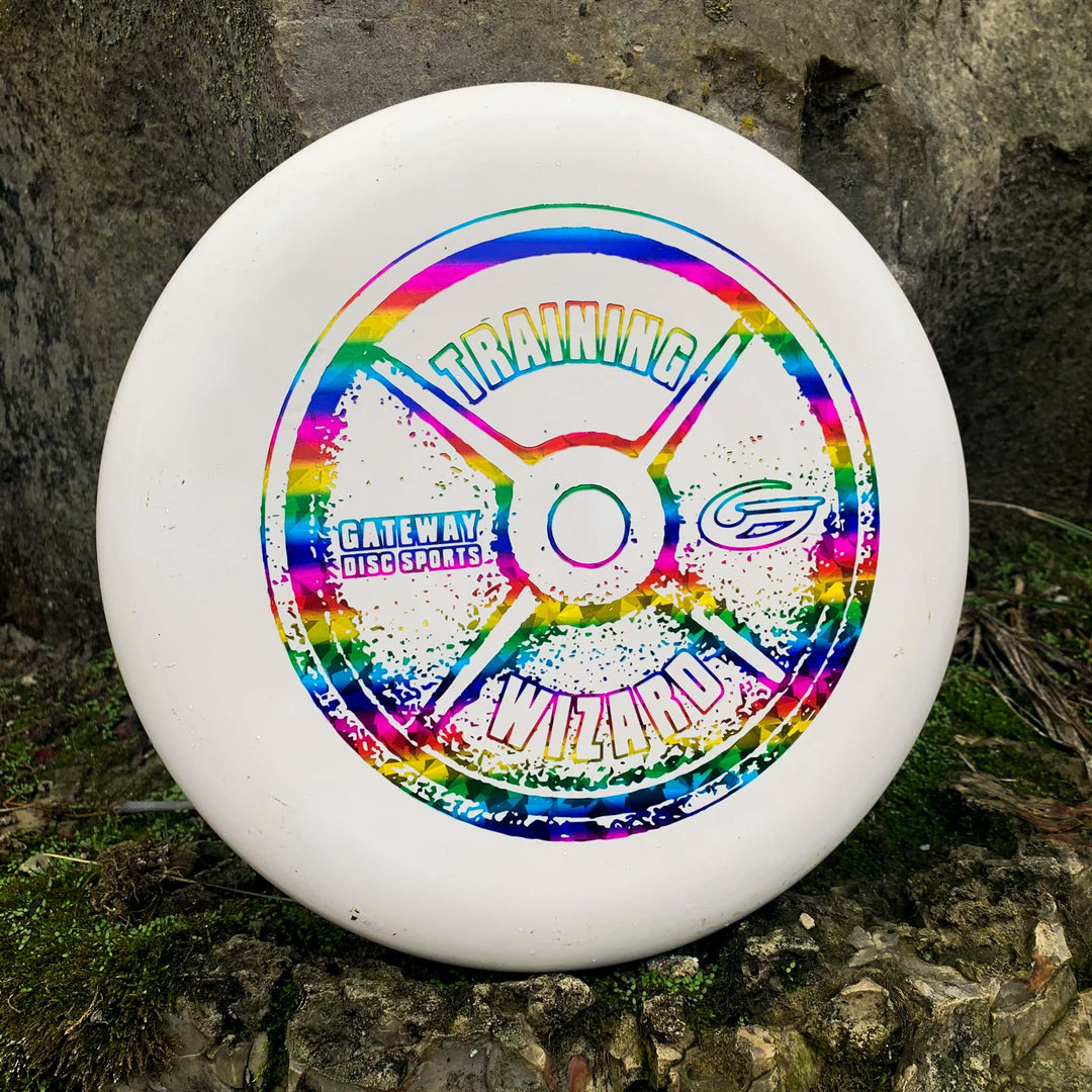 The Wizard - Training Weight is a white disc golf putter with a colorful "Training Wizard" design and Gateway Disc Sports logo, ideal as a stable workhorse for mid-range drives, shown resting on mossy ground against a rock.
