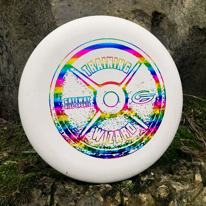 The Wizard - Training Weight is a white disc golf putter with a colorful "Training Wizard" design and Gateway Disc Sports logo, ideal as a stable workhorse for mid-range drives, shown resting on mossy ground against a rock.