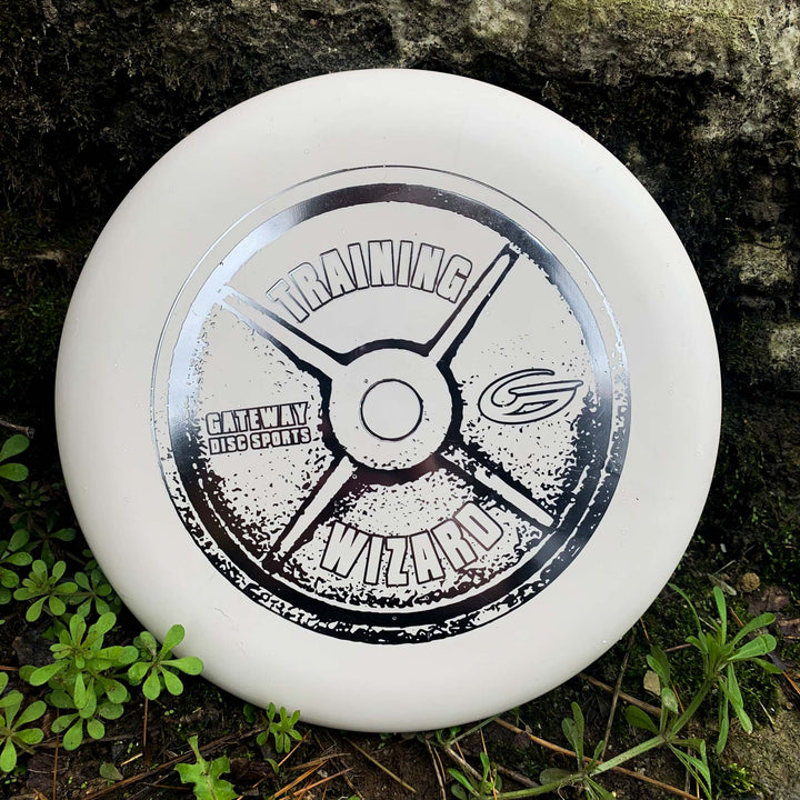 A white Gateway Wizard - Training Weight disc golf putter rests near green plants and a mossy rock, ready to be your stable putter of choice—even in windy conditions.