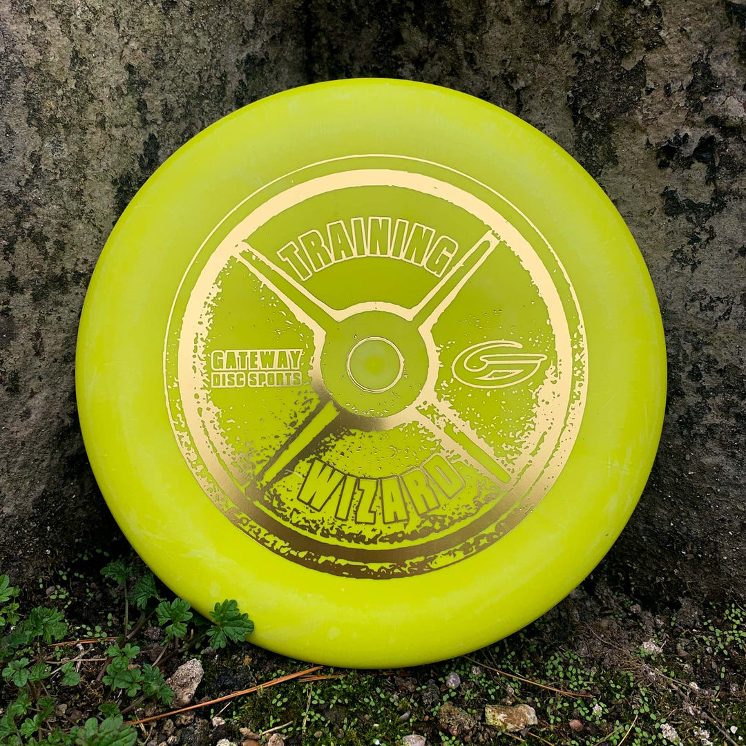 A yellow Gateway Disc Sports "Wizard - Training Weight" disc golf putter, a stable workhorse for mid-range drives, stands upright against a tree trunk with grass and moss at the base.