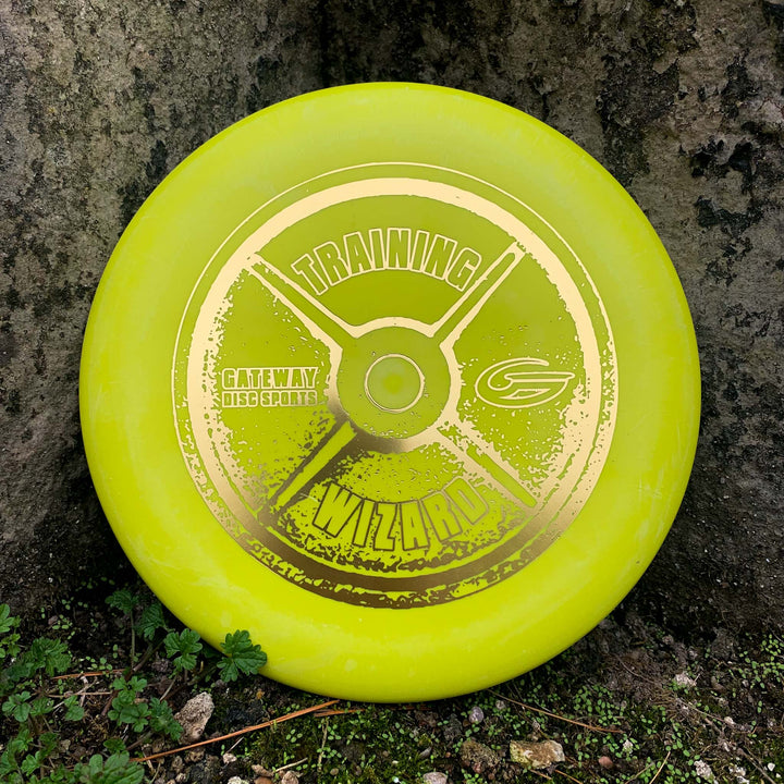 A yellow Gateway Disc Sports "Wizard - Training Weight" disc golf putter, a stable workhorse for mid-range drives, stands upright against a tree trunk with grass and moss at the base.