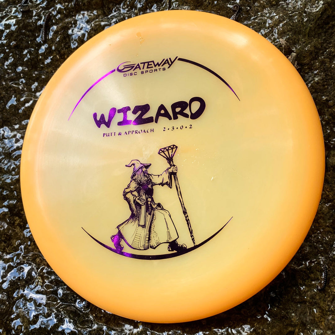 A yellow Wizard - Diamond putt and approach disc, featuring a wizard illustration, is shown resting on a wet, textured surface.