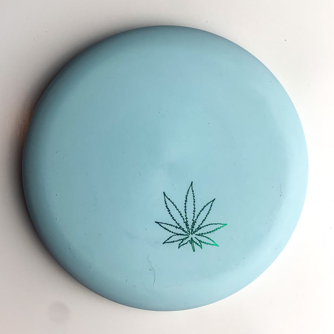 A light blue Wizard Leaf Suregrip Putter from Gateway Disc Sports, featuring an iconic leaf design near the edge, sits on a white surface—ideal for fans of Wizard putters.