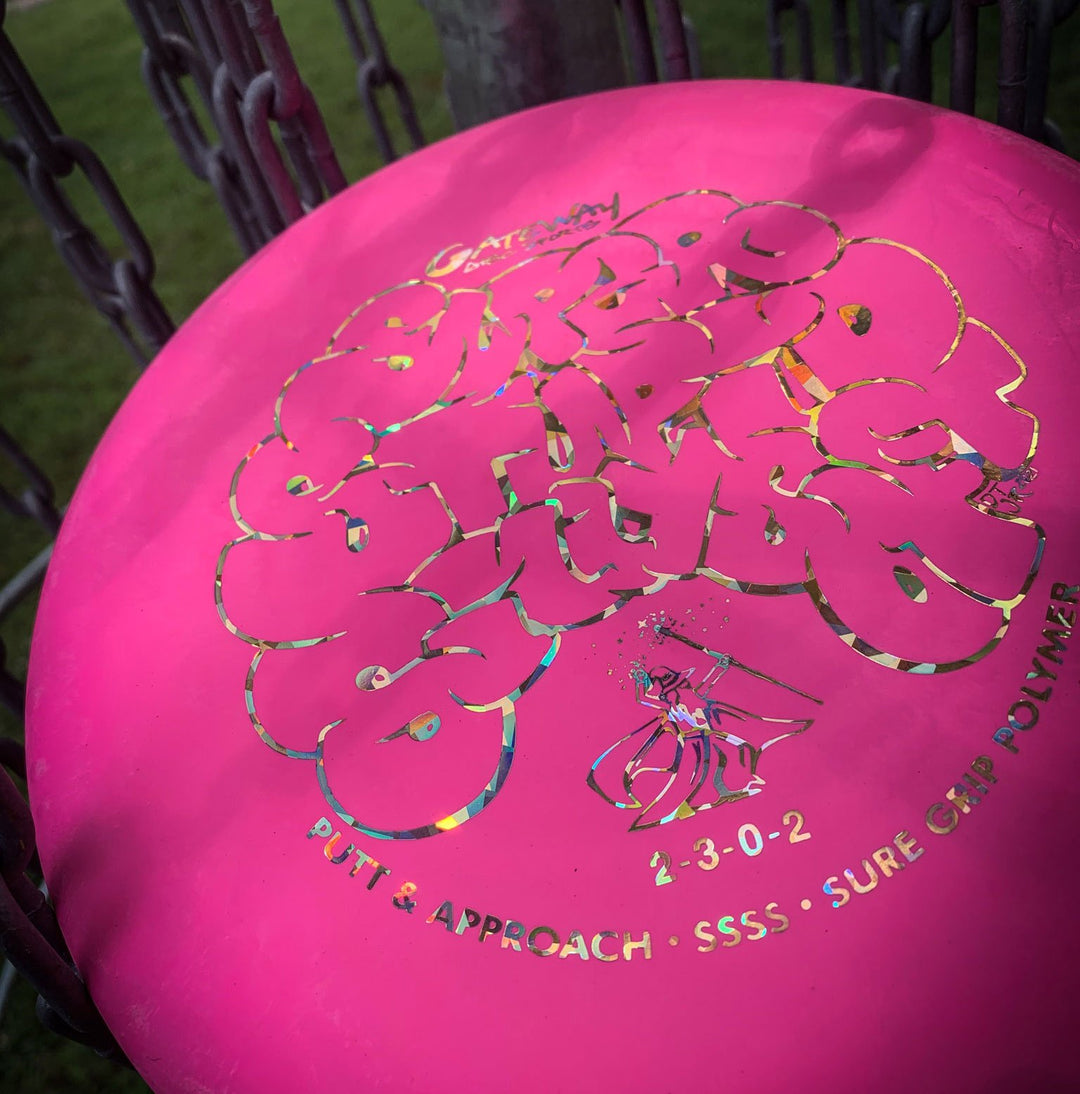 A pink Wizard - SSSS putt and approach disc with "Stupid Stupid Thumb" beading is shown resting in a metal basket, grass visible in the background.