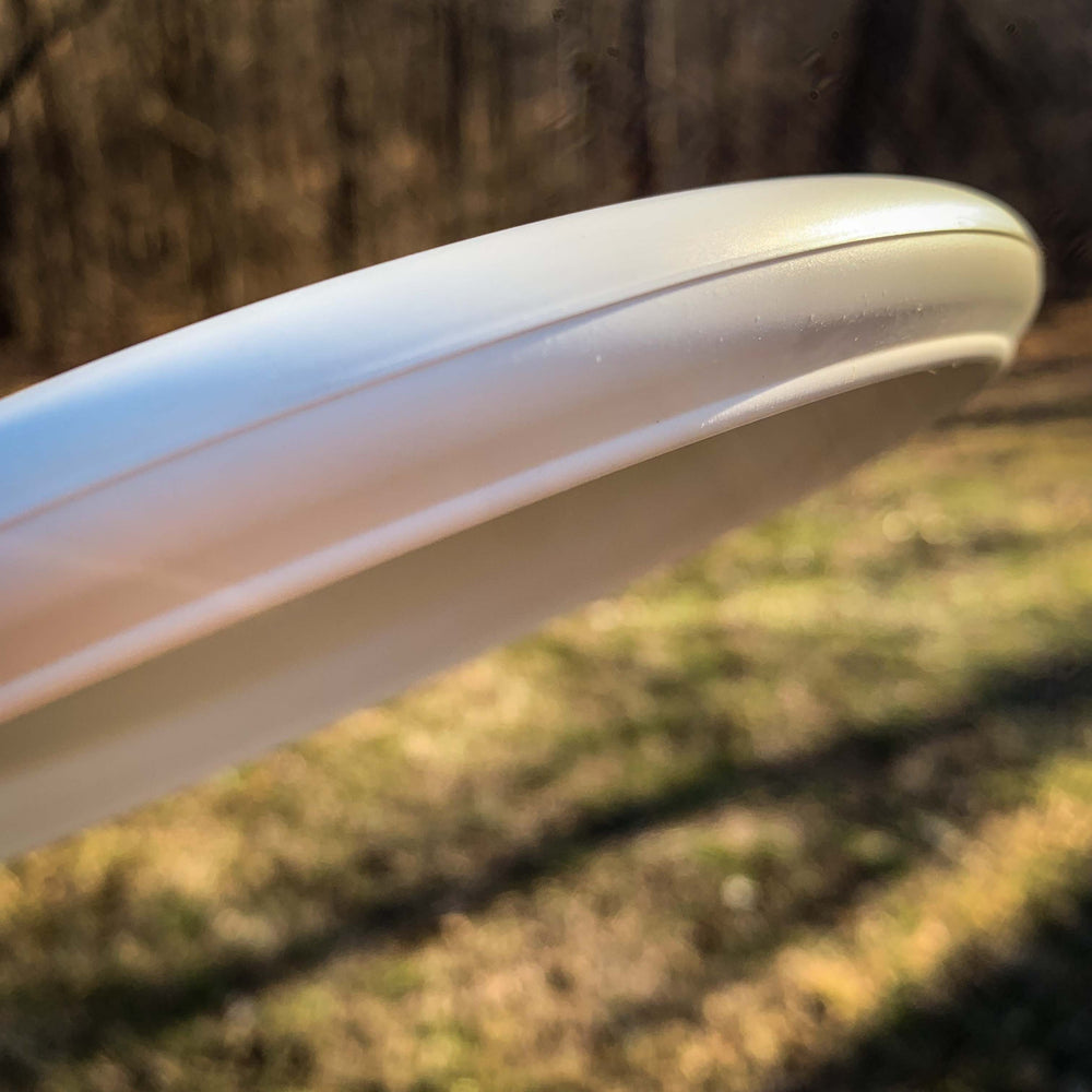 A close-up of the edge of a "Not So" Pure White Wizard disc held outdoors, with sunlit trees and grass in the background—ideal as a stable workhorse putter for controlled mid-range drives.
